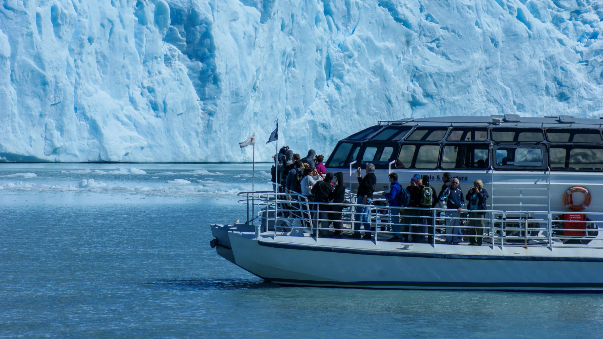 A small excursion boat with passengers on its bow rail close to a Patagonian glacier ice wall